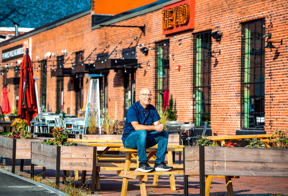A smiling white man sits on a picnic table on a brick patio in front of a long brick building with neon signs