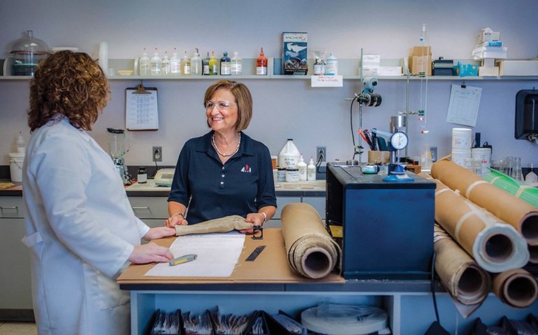 A woman wearing safety glasses talks to another woman in a lab coat at a table with rolled-up textiles.