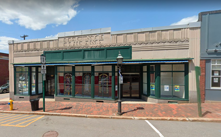 A one story art deco building on a street corner that looks vacant with a green sign thats blank