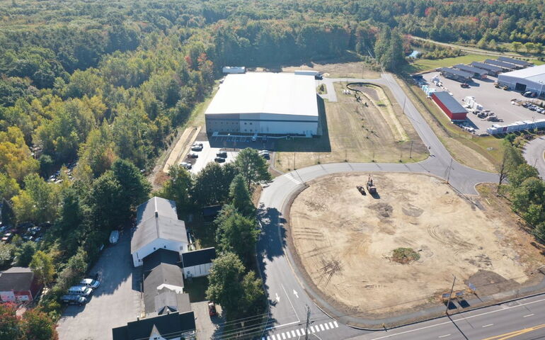 An aerial view of buildings, woods and an empty lot.