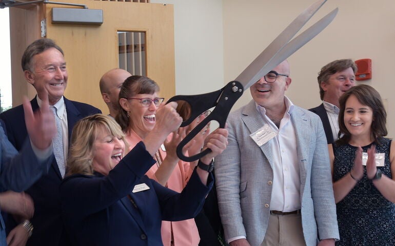 A group of people are smiling and clapping while one holds oversize scissors.