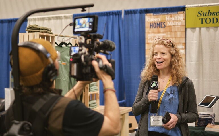 A person speaks into a microphone while another person holds a camera.