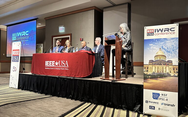 People sit and stand on a stage at a lectern and long table.