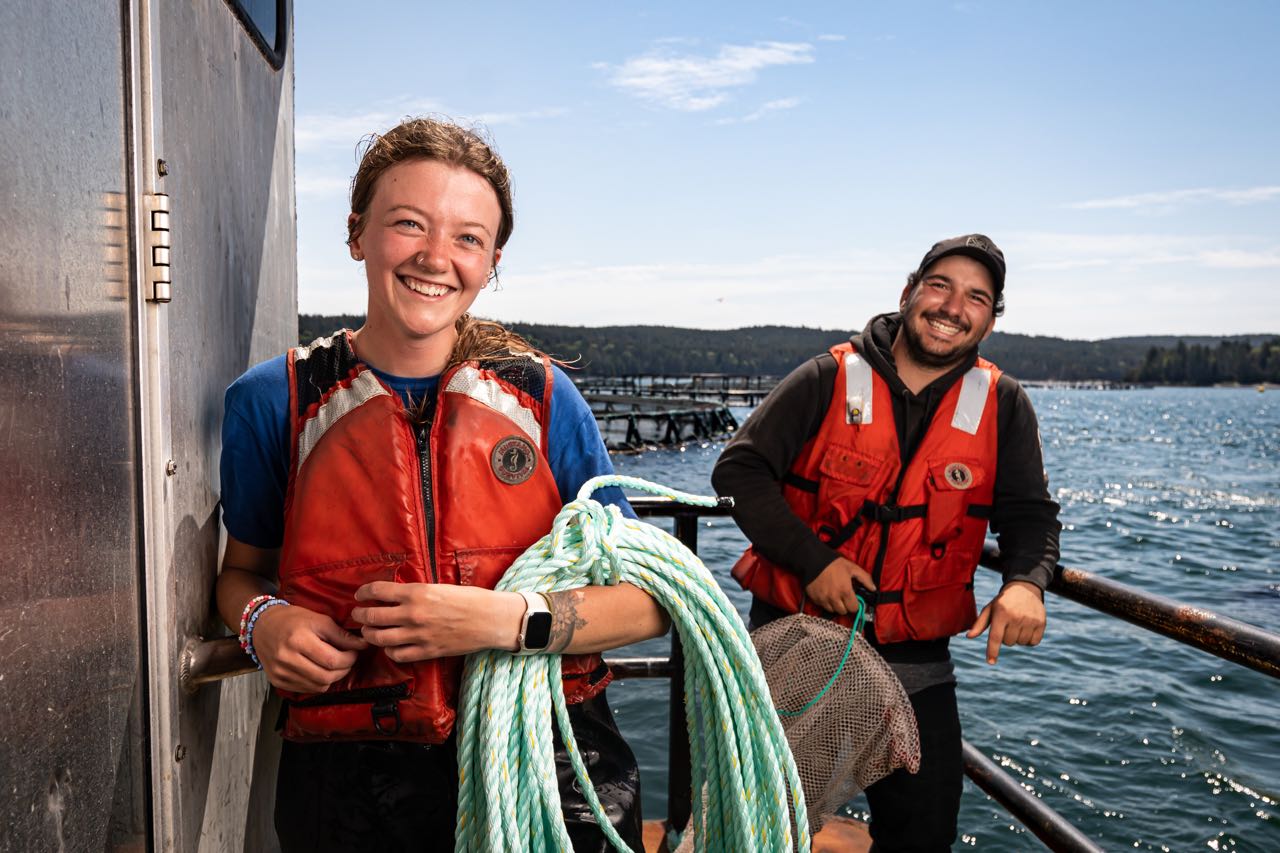 Two people stand on a barge, one holding a coil of rope.