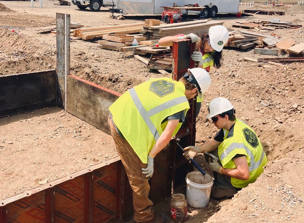 Three students with the Maine Construction Academy learn how to build a foundation. They're looking at tools next to some steel set in the ground.