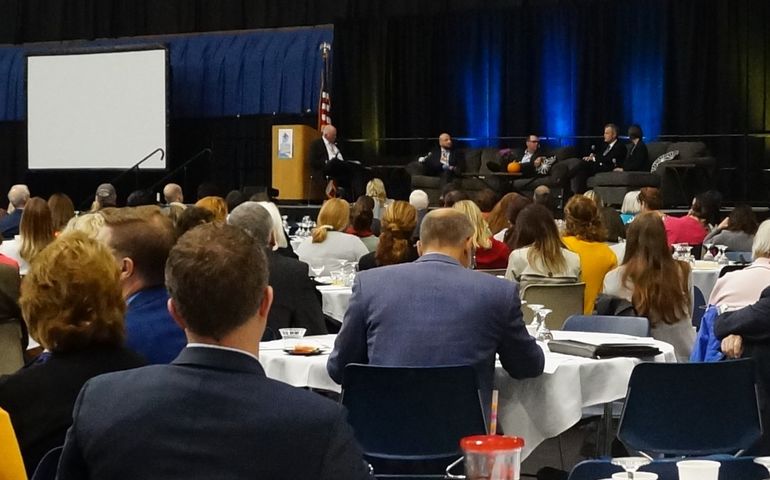 a crowd in a conference hall with a panel of five people way in the background on a stage