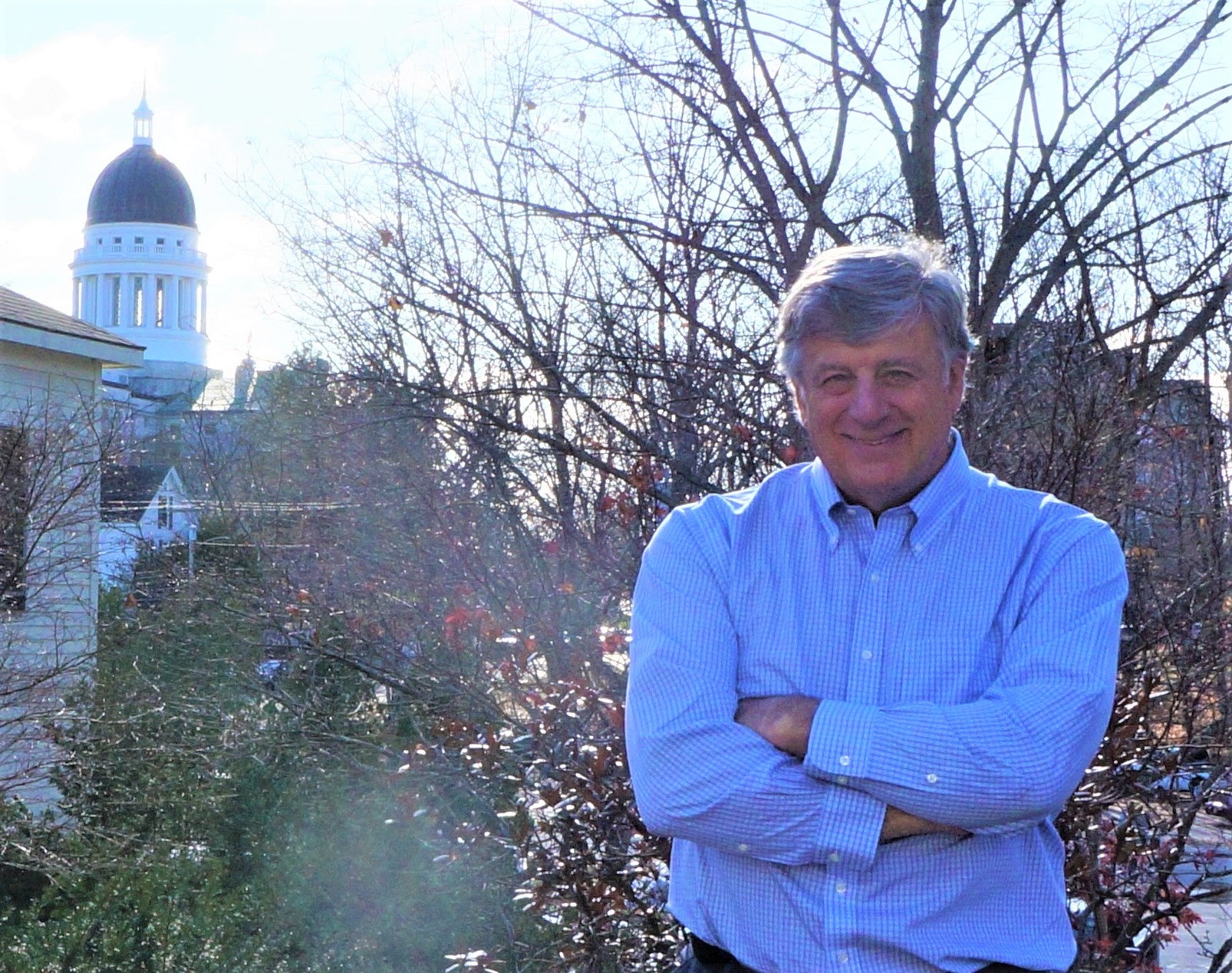 A man stands outdoors with his arms folded and the dome of the Maine State House in the backround