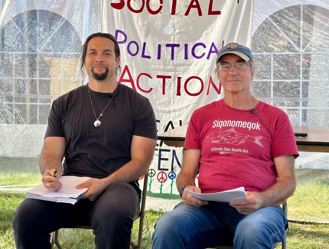 Two people sit with documents under a tent.