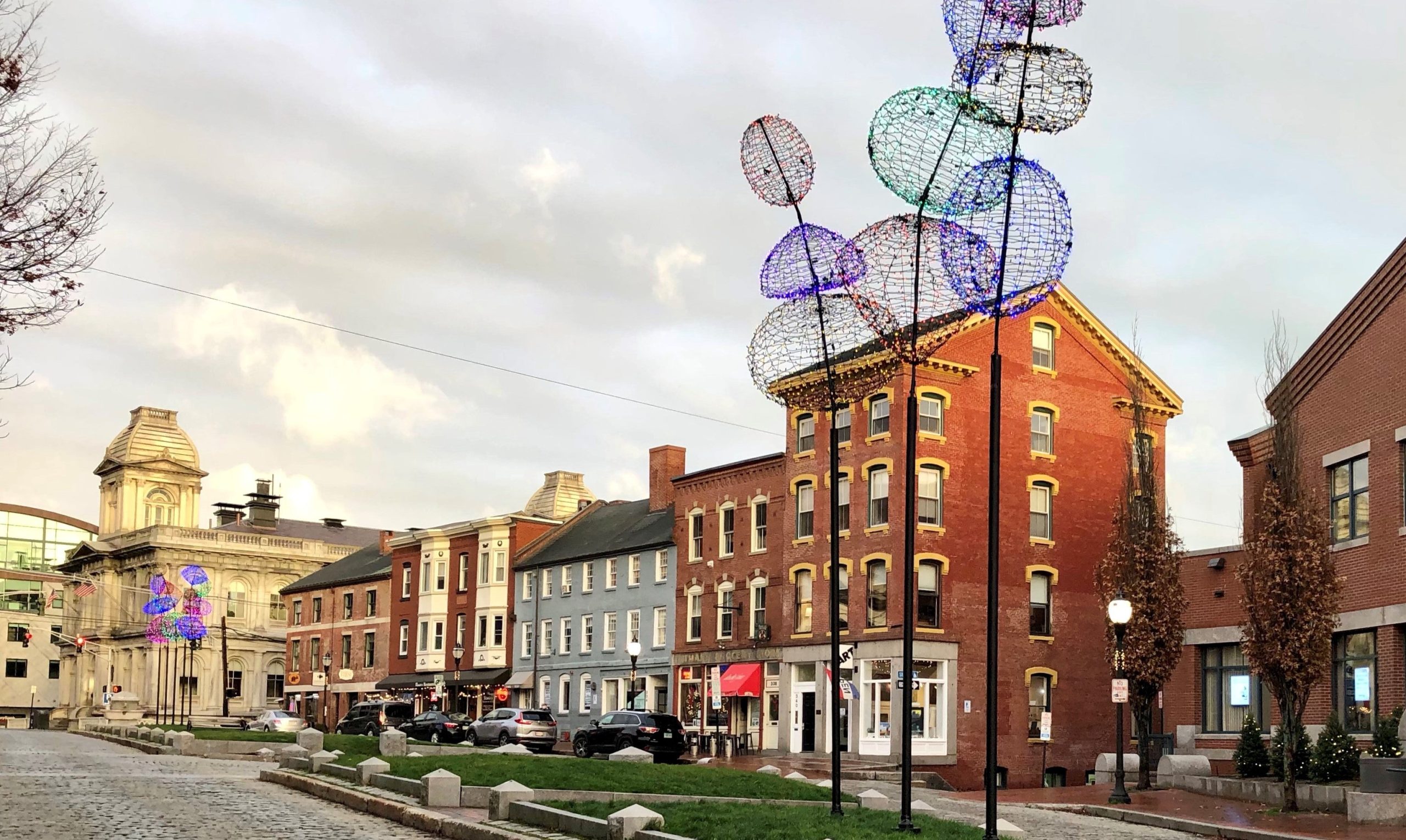 A cobblestone street with a variety of brick and brightly painted buildings a and a grassy median with granite curbing