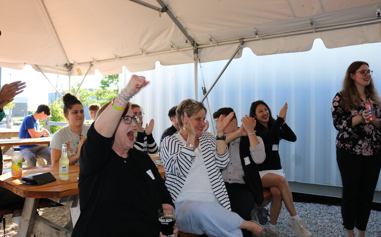 A group of people sit at picnic tables under a tent and applaud.