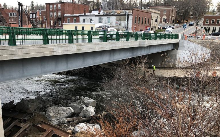 a concrete bridge over a rocky stream with brick two and three story buildings on the other side