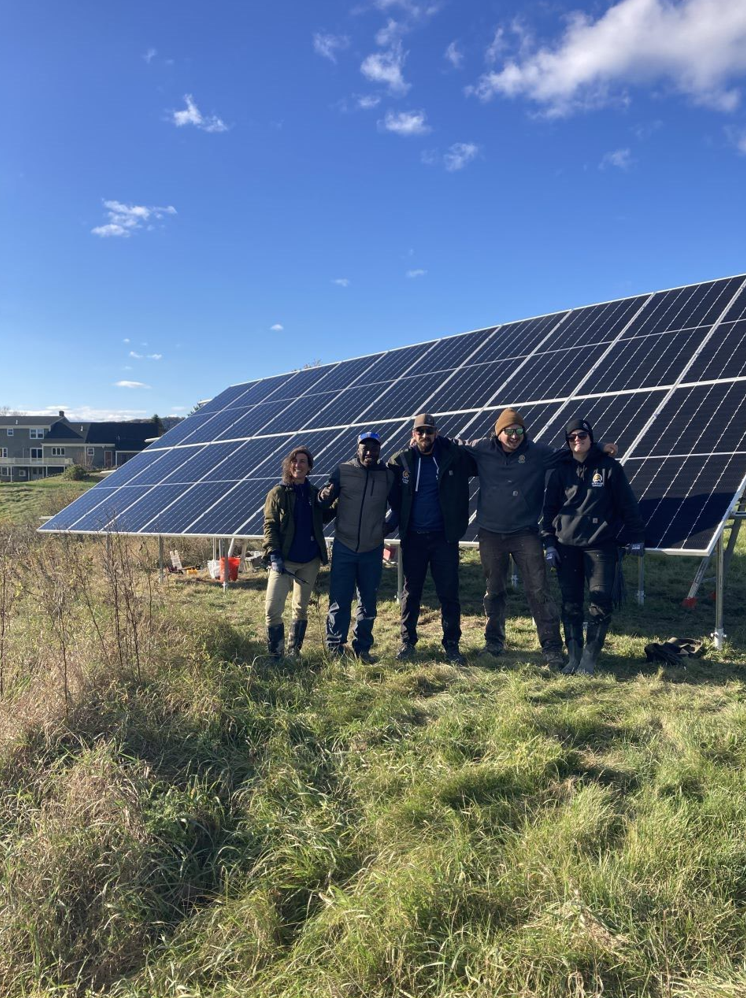 Five people stand in a field in front of solar panels.