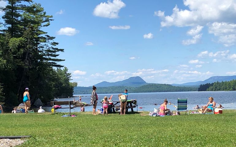 a grassy beach with people in swimsuits and a blue lake beyond and mountains beyond that