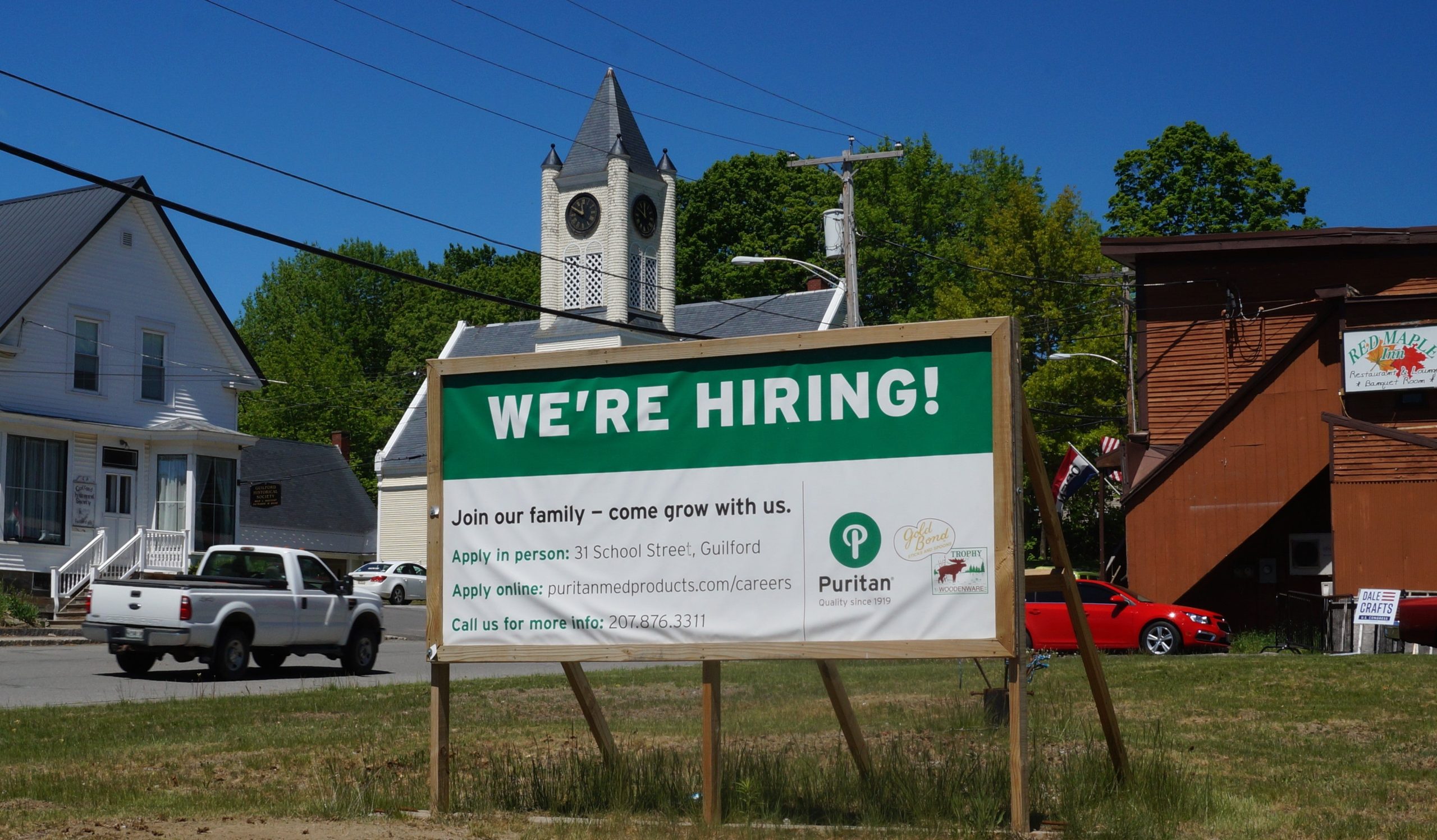 A sign that says we're hiring puritan medical products on a street with a wooden church and other small maine town buildings