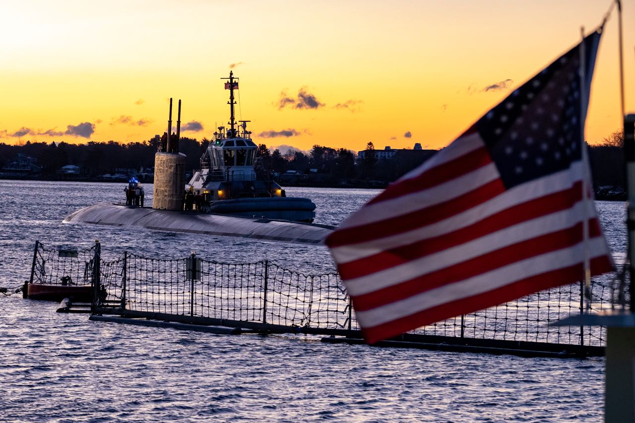 A flag flies in the foreground and a sub is in the water in the background.